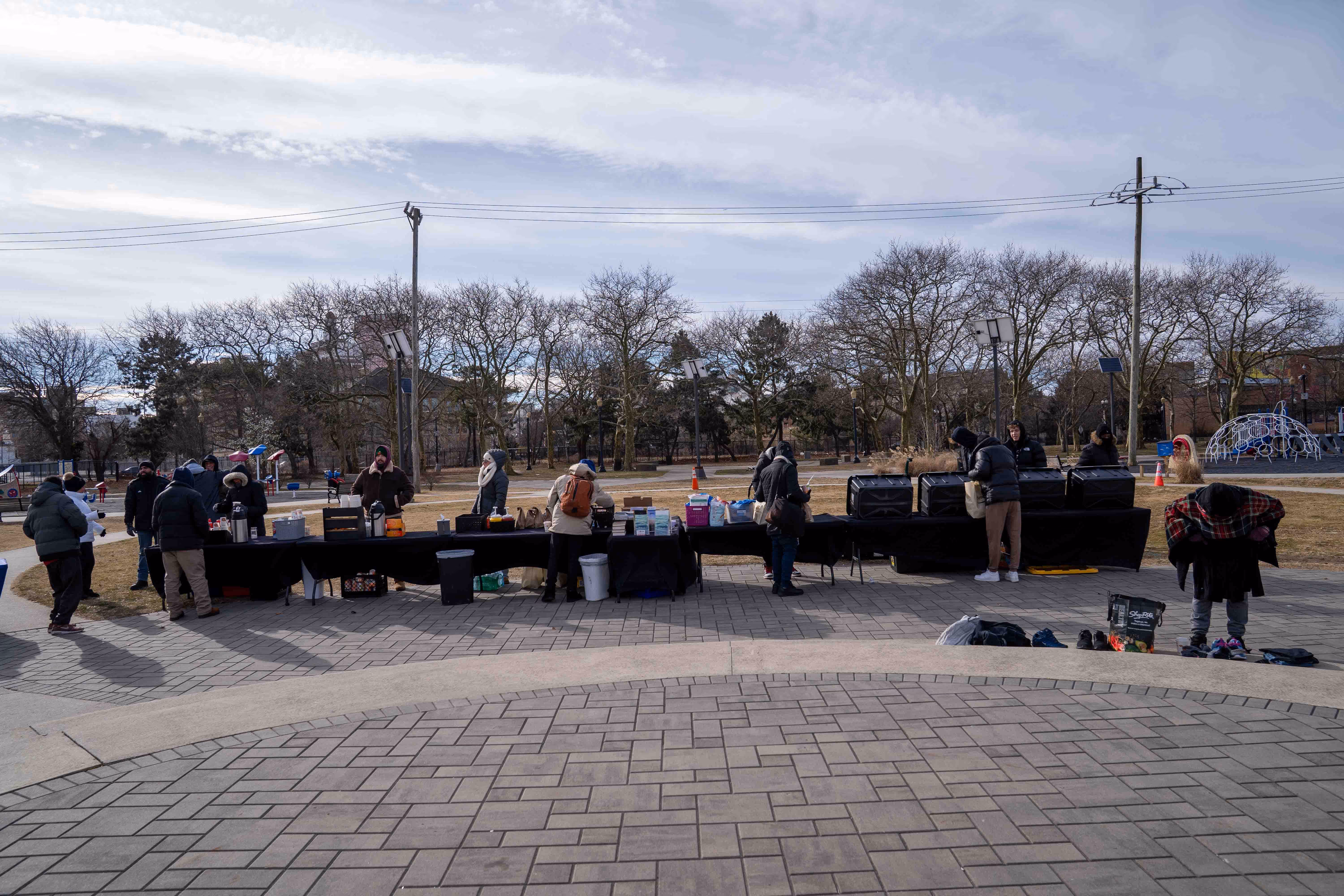 People bundled in winter clothes standing and serving at a long outdoor table with food and supplies in a park.