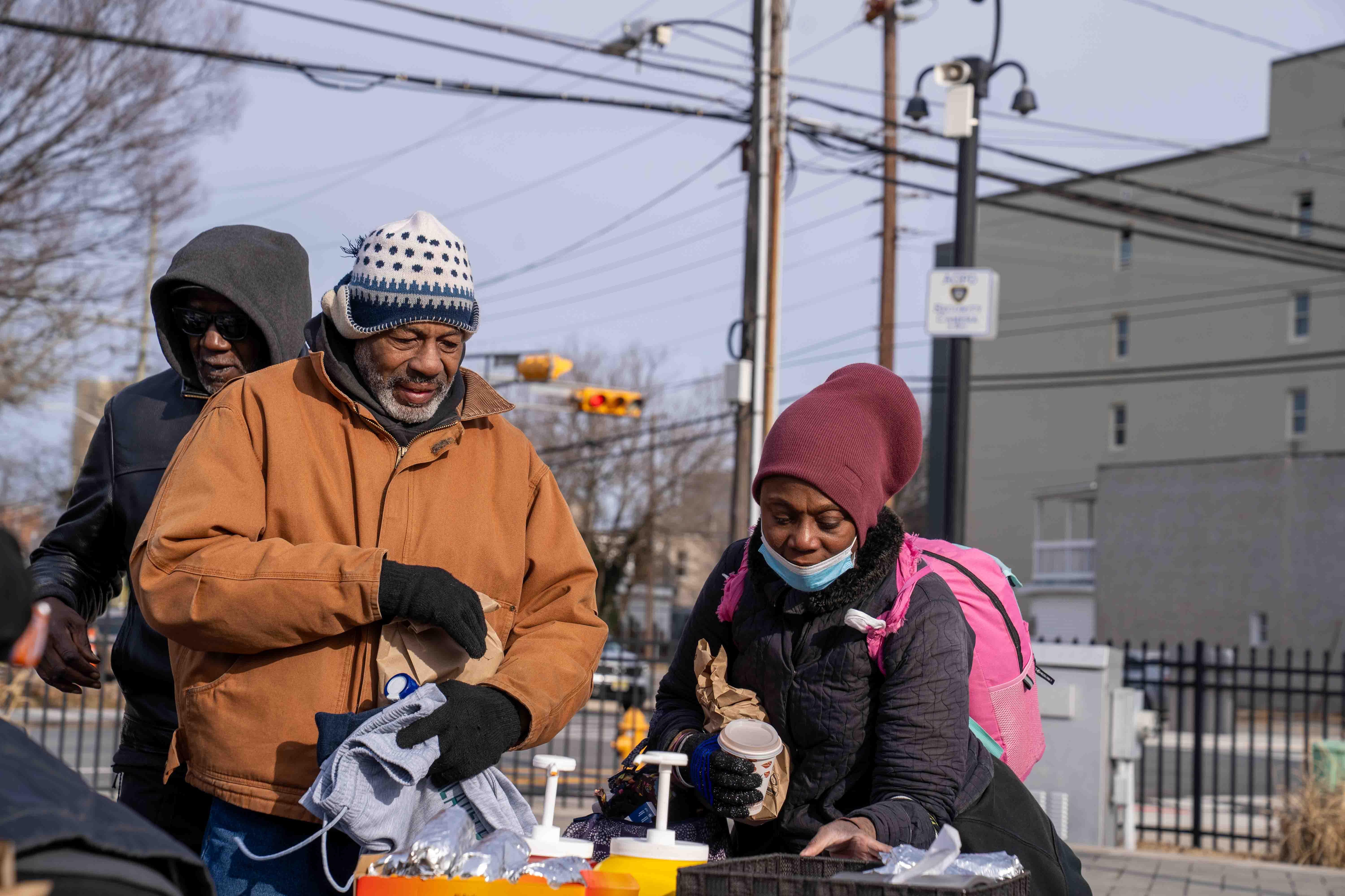 Three people in winter clothing, one holding a takeaway coffee cup, gathered outdoors near a table with foil-wrapped items and bottles with pumps.