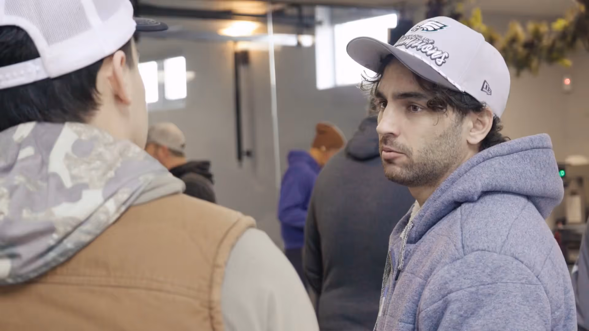 Two men wearing hats and hoodies engaged in a conversation indoors with other people in the background.