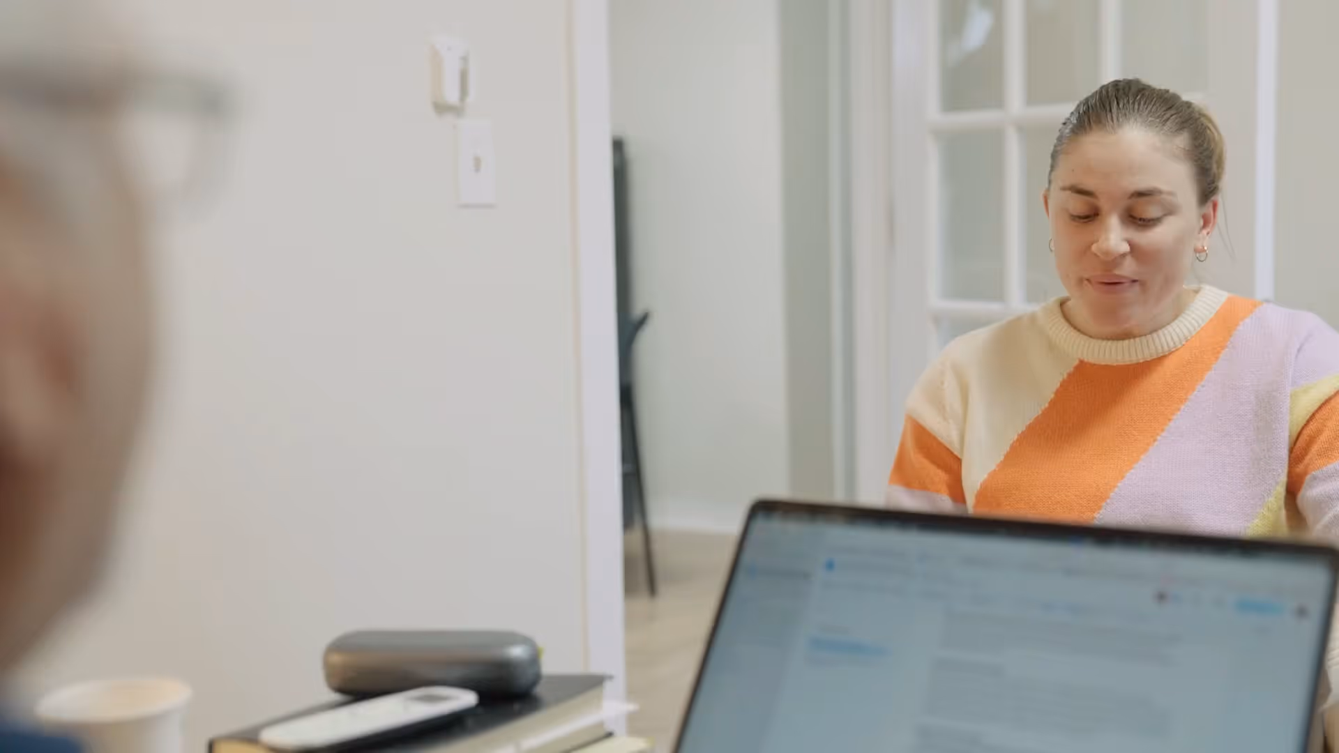 Person in orange and purple sweater talking to someone using a laptop in a bright room.