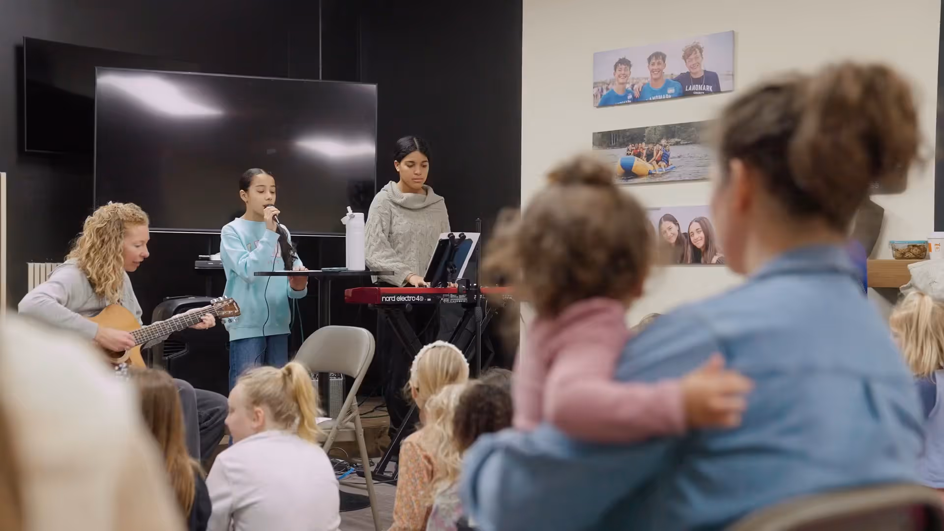 Woman plays guitar, girl sings with microphone, and another plays keyboard in front of seated children.
