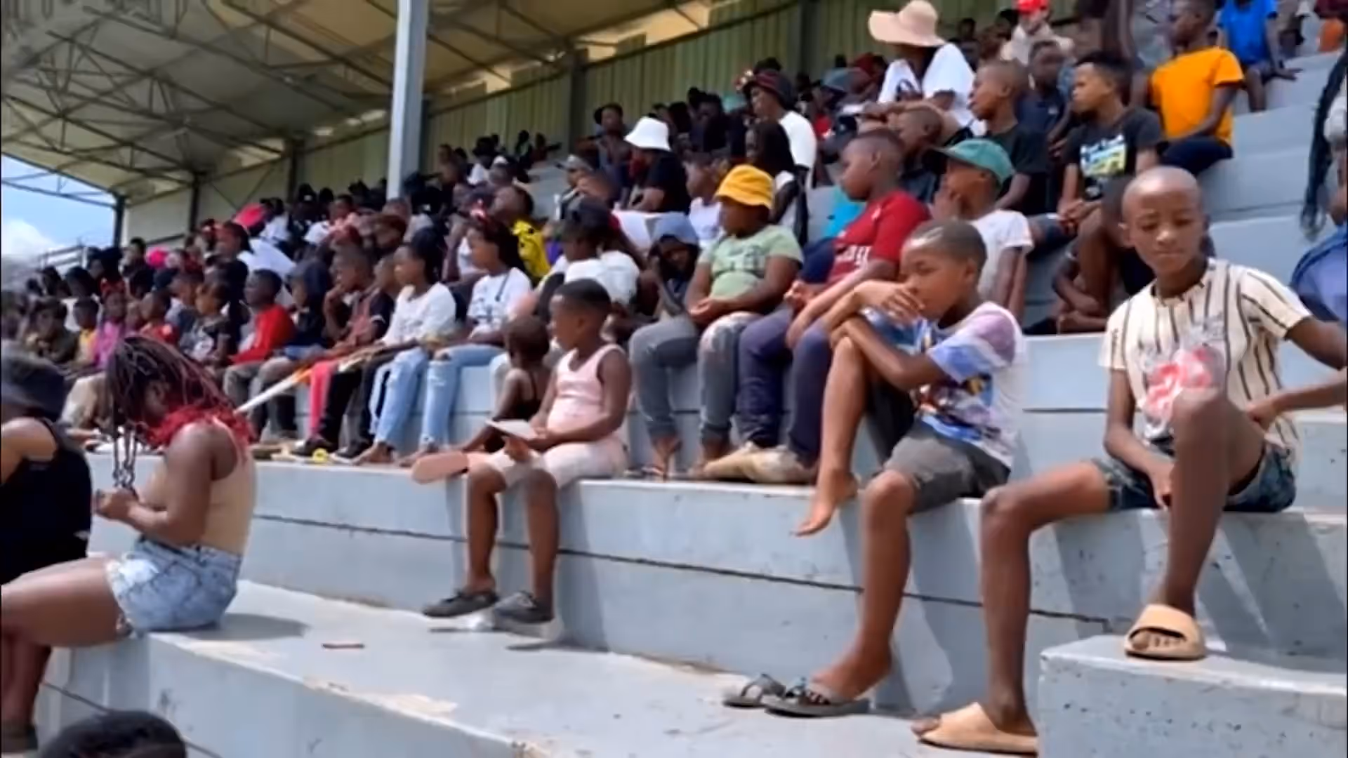 A large group of children and teens sitting on concrete bleachers under a covered area.
