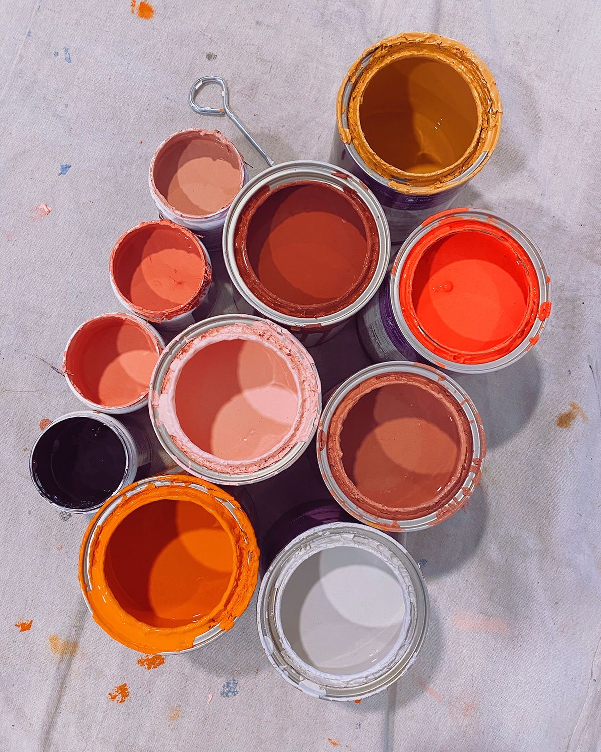 Several open paint cans containing various warm colors including orange, red, brown, pink, and white arranged on a cloth drop cloth.