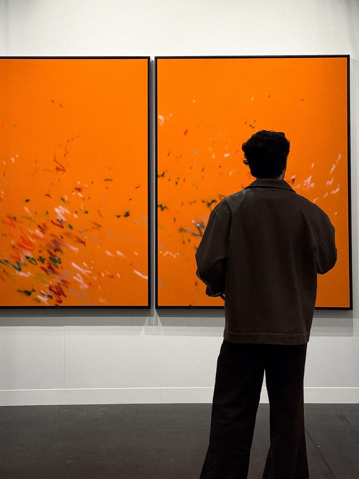 Person with dark hair and jacket standing in front of two large orange abstract paintings with scattered colorful brush strokes in an art gallery.