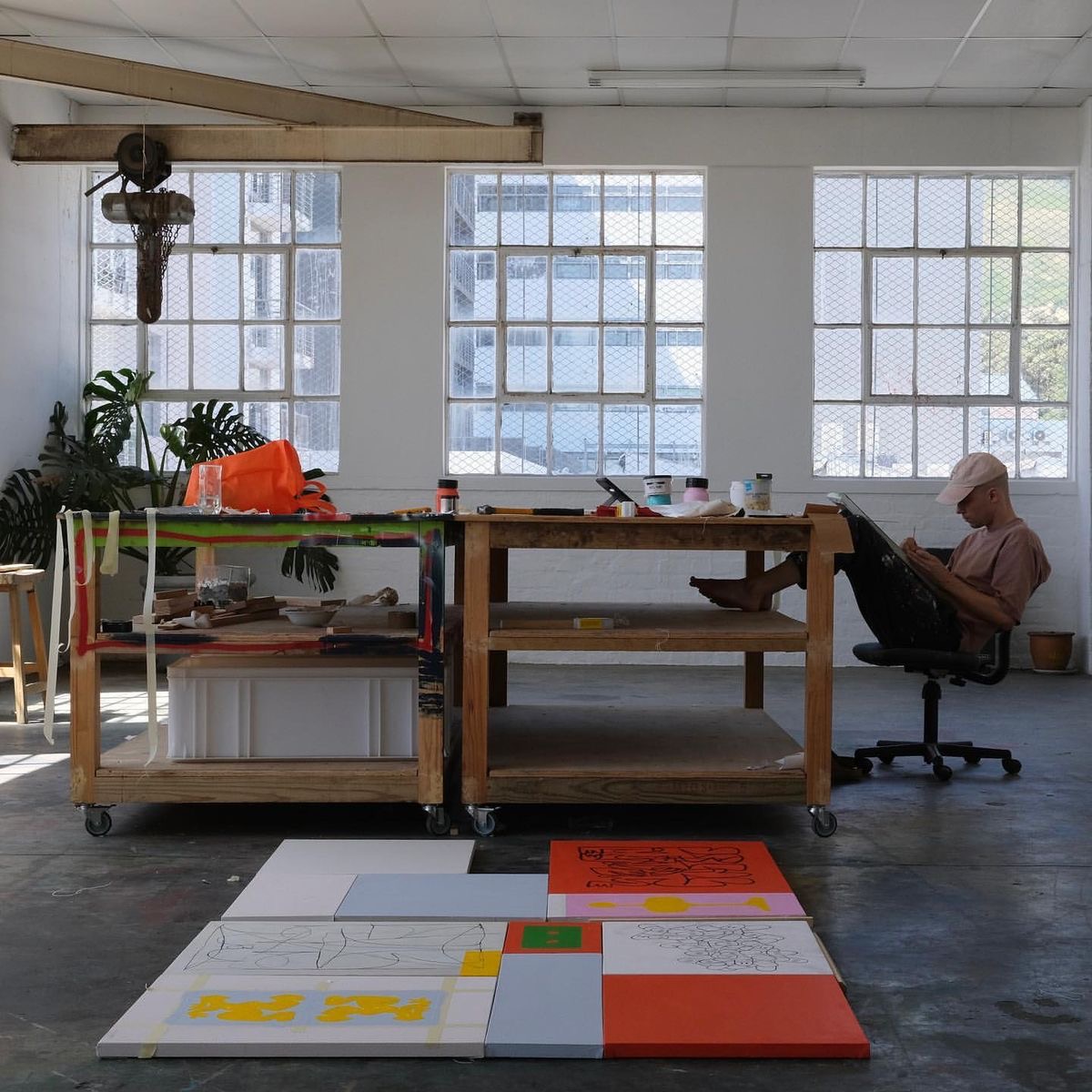 Artist sketching while seated on a black rolling chair next to a wooden workbench with art supplies, with several colorful artworks laid out on the floor in front.