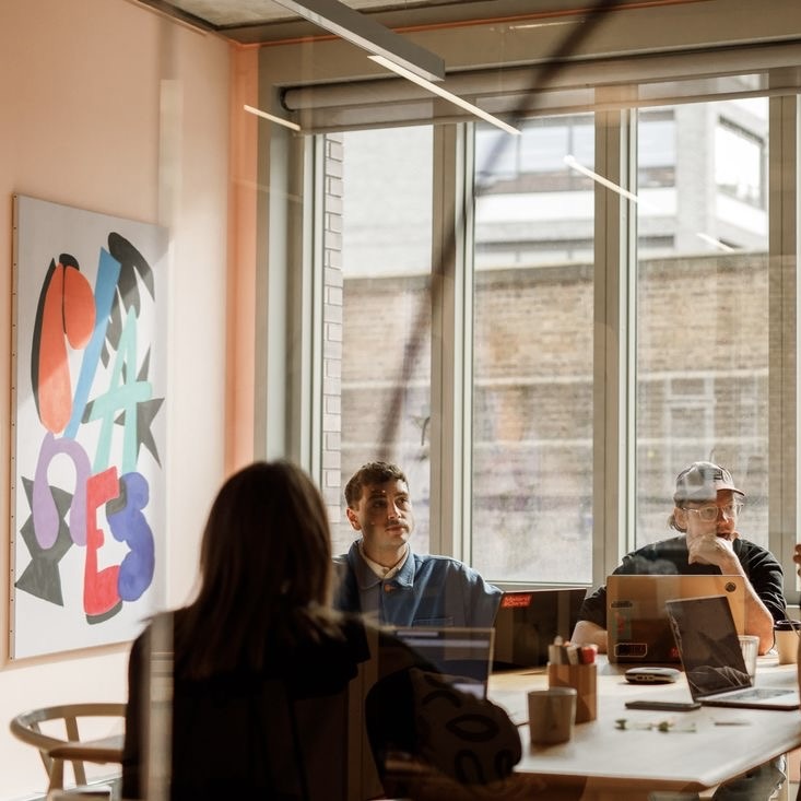 Three people working on laptops around a wooden table in a well-lit office with large windows and abstract colorful wall art.