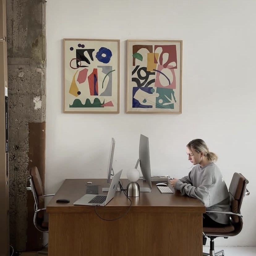 Woman sitting at a wooden desk with dual monitors and a laptop, looking at her phone in a minimalist office with abstract framed art on the wall.