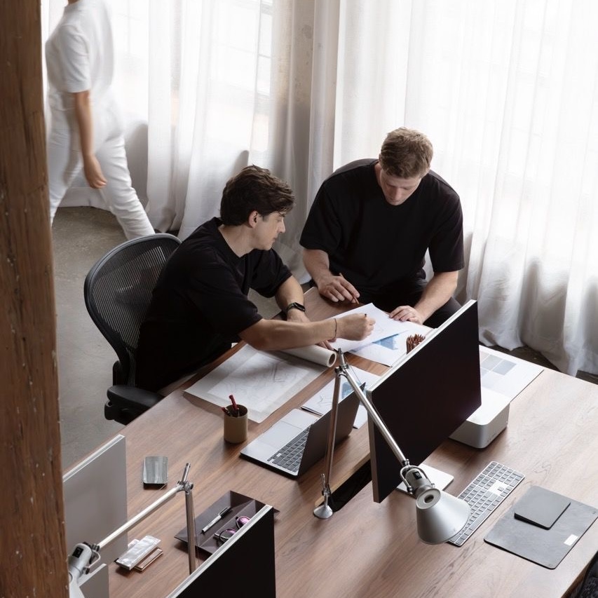 Two men in black shirts working on drawings and discussing at a desk with laptops and monitors in a bright office while a person in white walks by.