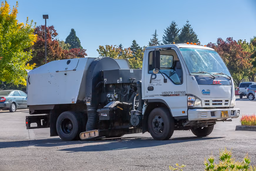 PMI Paving branded parking lot sweeper truck parked in a lot with autumn trees