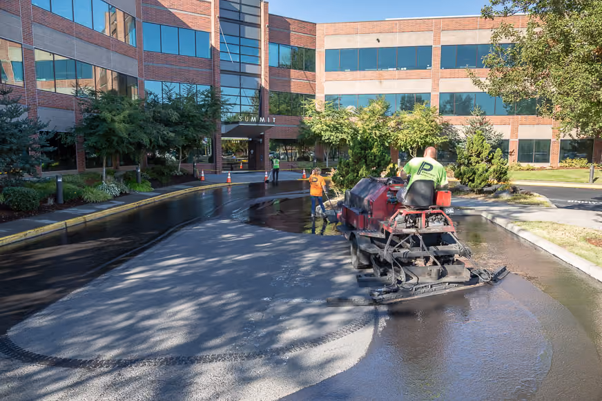 PMI Paving crew member sealcoating the entrance drive of a brick office building, illustrating the Property/Facility Managers industry card in the homepage's "Who we serve" section