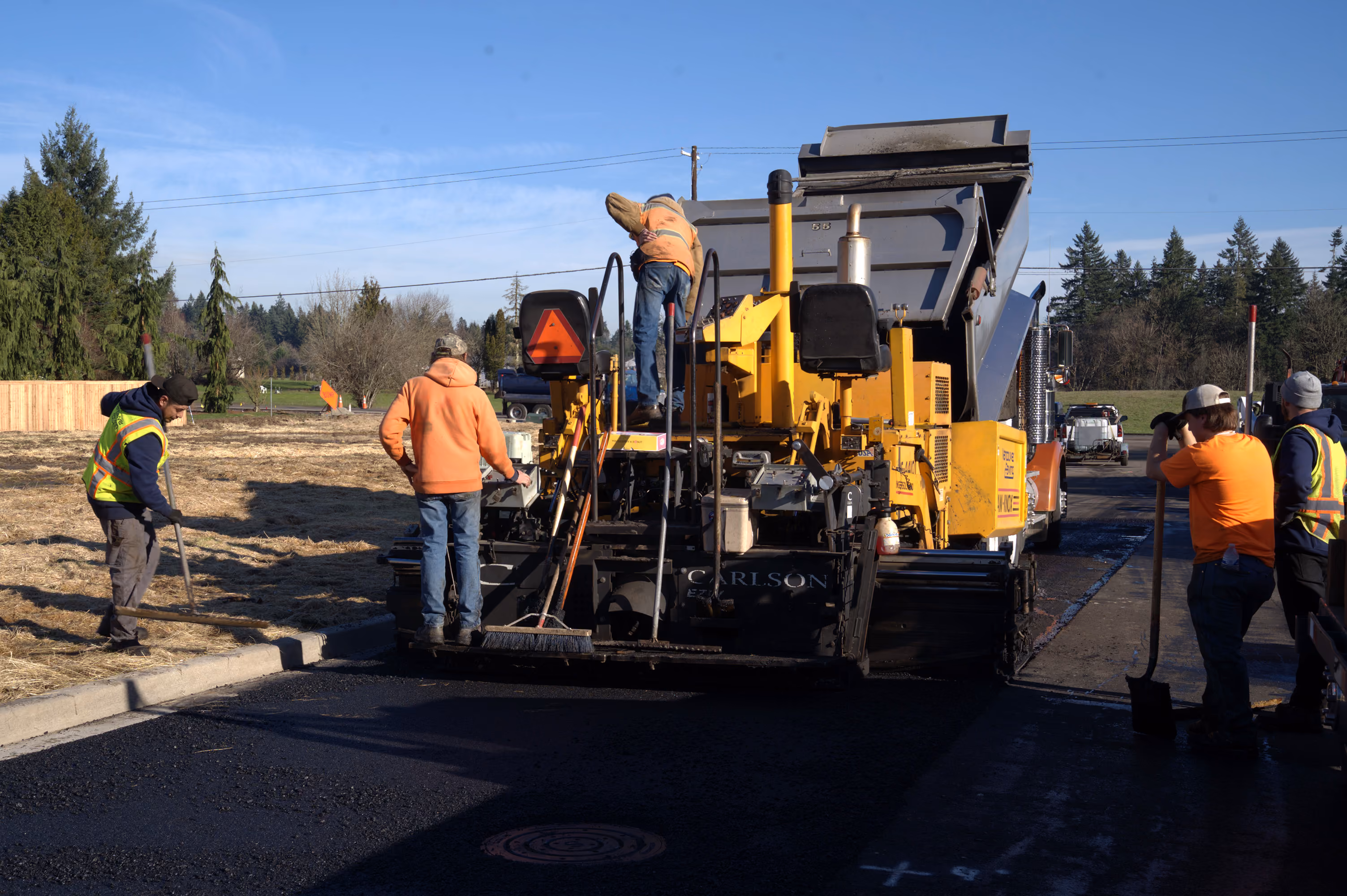 Wide shot of a PMI Paving crew laying fresh asphalt with a paver machine on a street, several workers in orange safety vests, trees lining the road in the background
