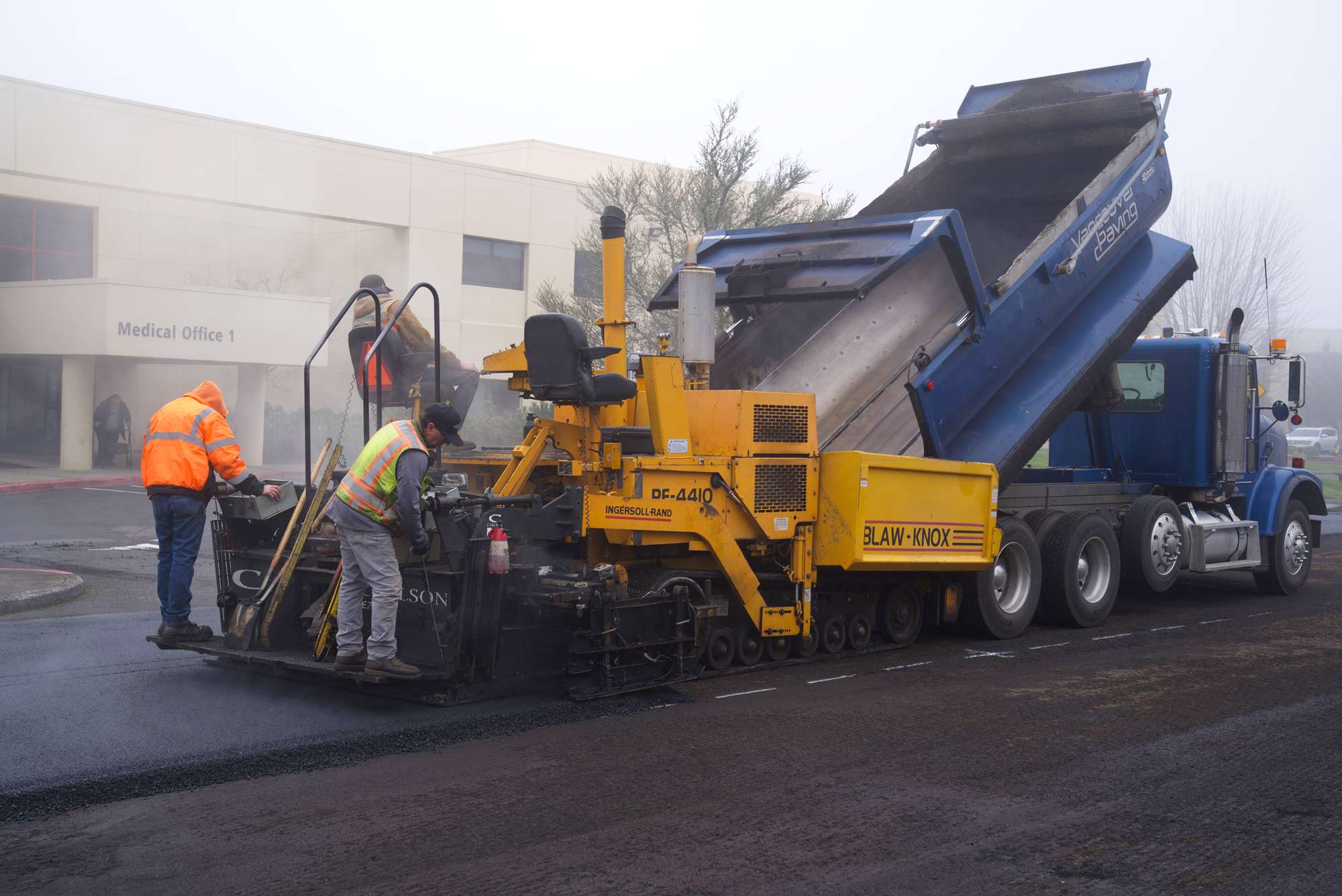 PMI Paving crew loading an asphalt paver from a dump truck in front of a "Medical Office 1" building on a foggy morning