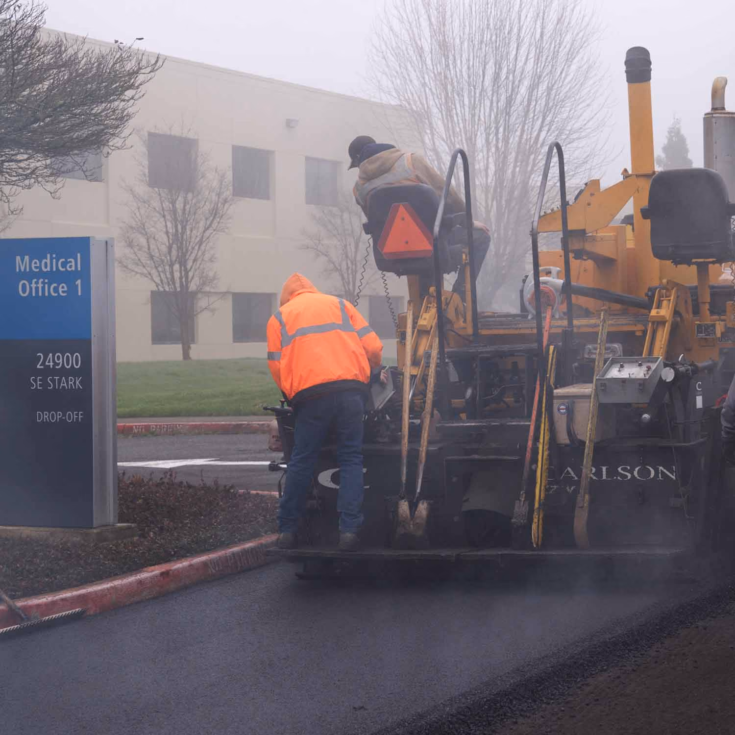 PMI Paving crew operating an asphalt paver in front of a "Medical Office 1" building sign on a foggy morning