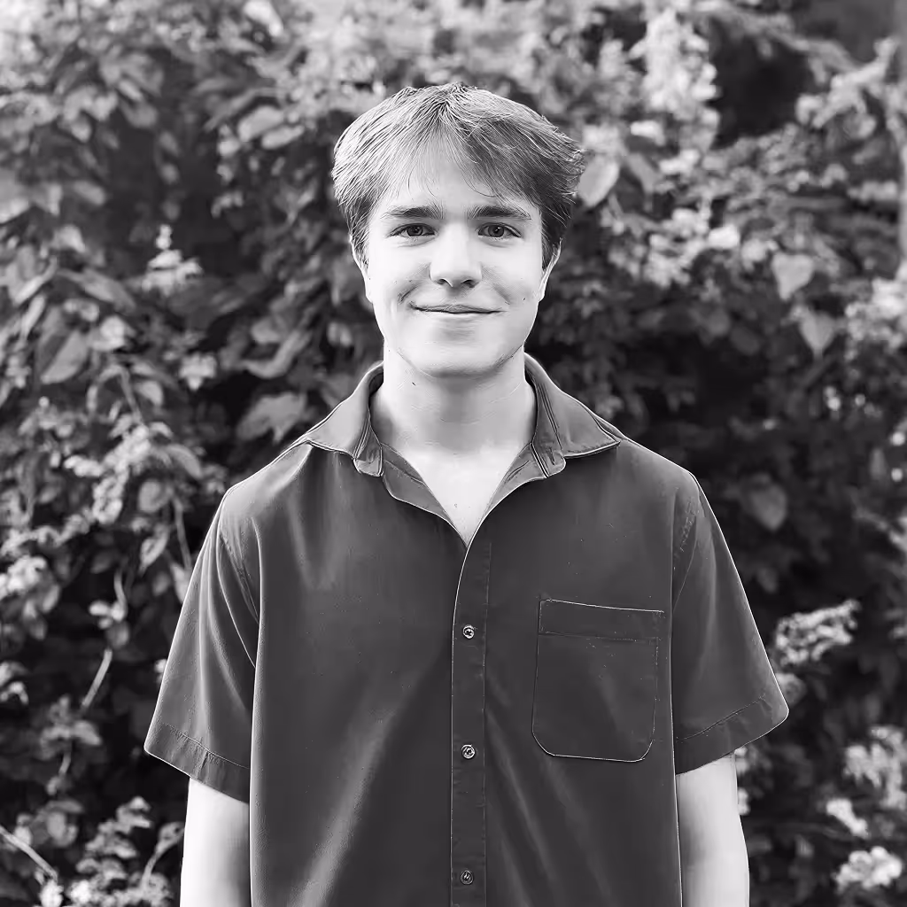 Young man with short hair smiling, wearing a short-sleeve buttoned shirt, standing in front of leafy background.