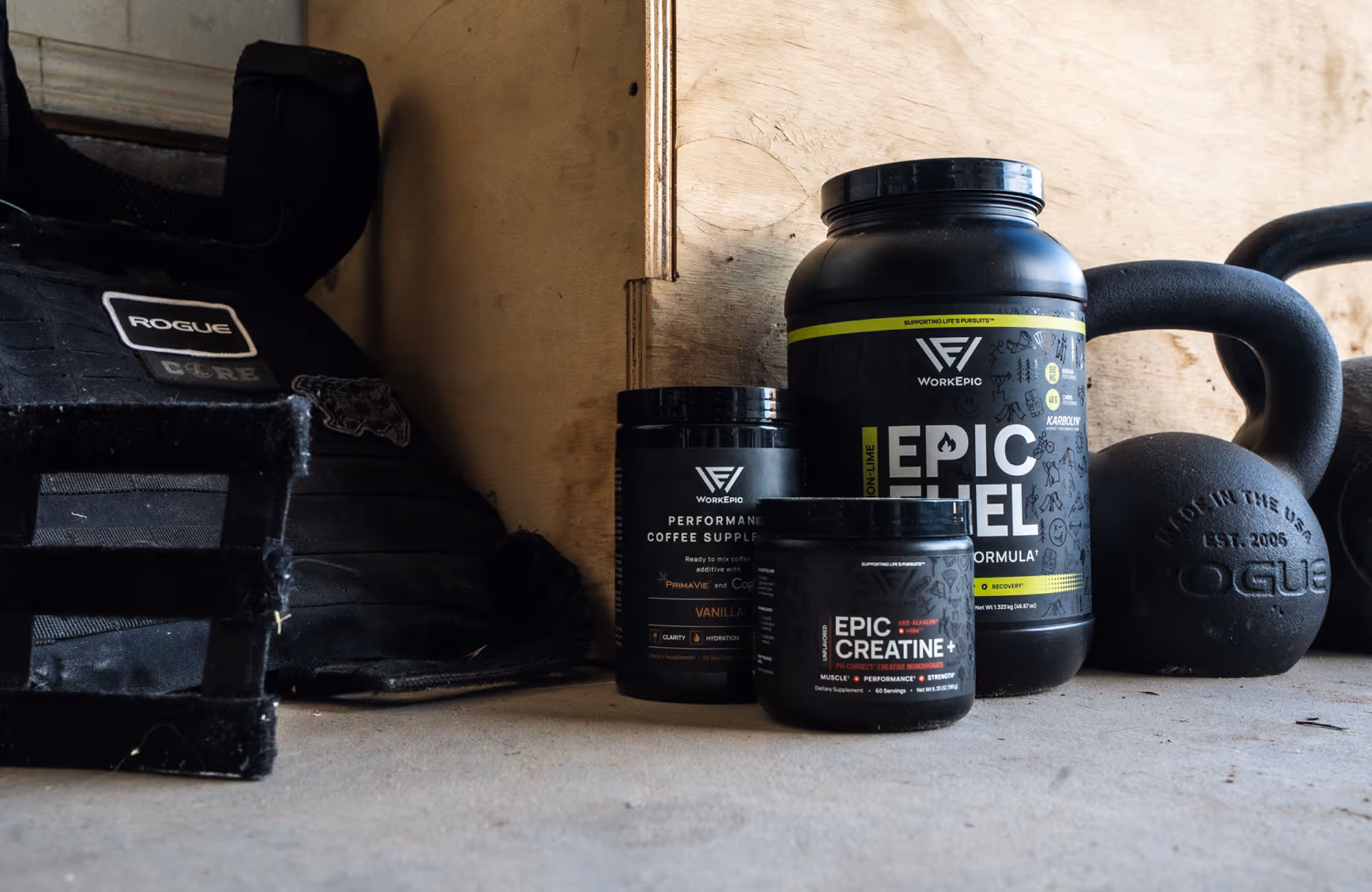 Three black WorkEpic supplement containers placed on a floor next to a kettlebell and Rogue weight vest in a workout space.