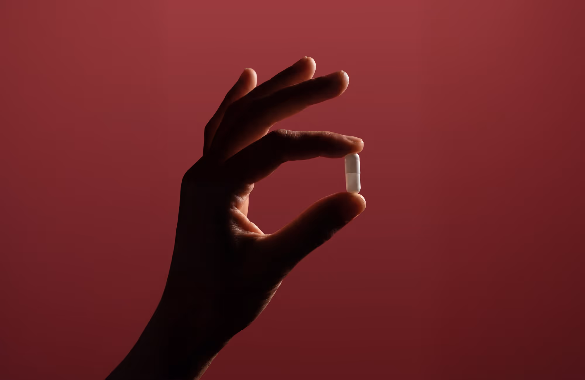 Hand holding a small white capsule pill against a dark red background.