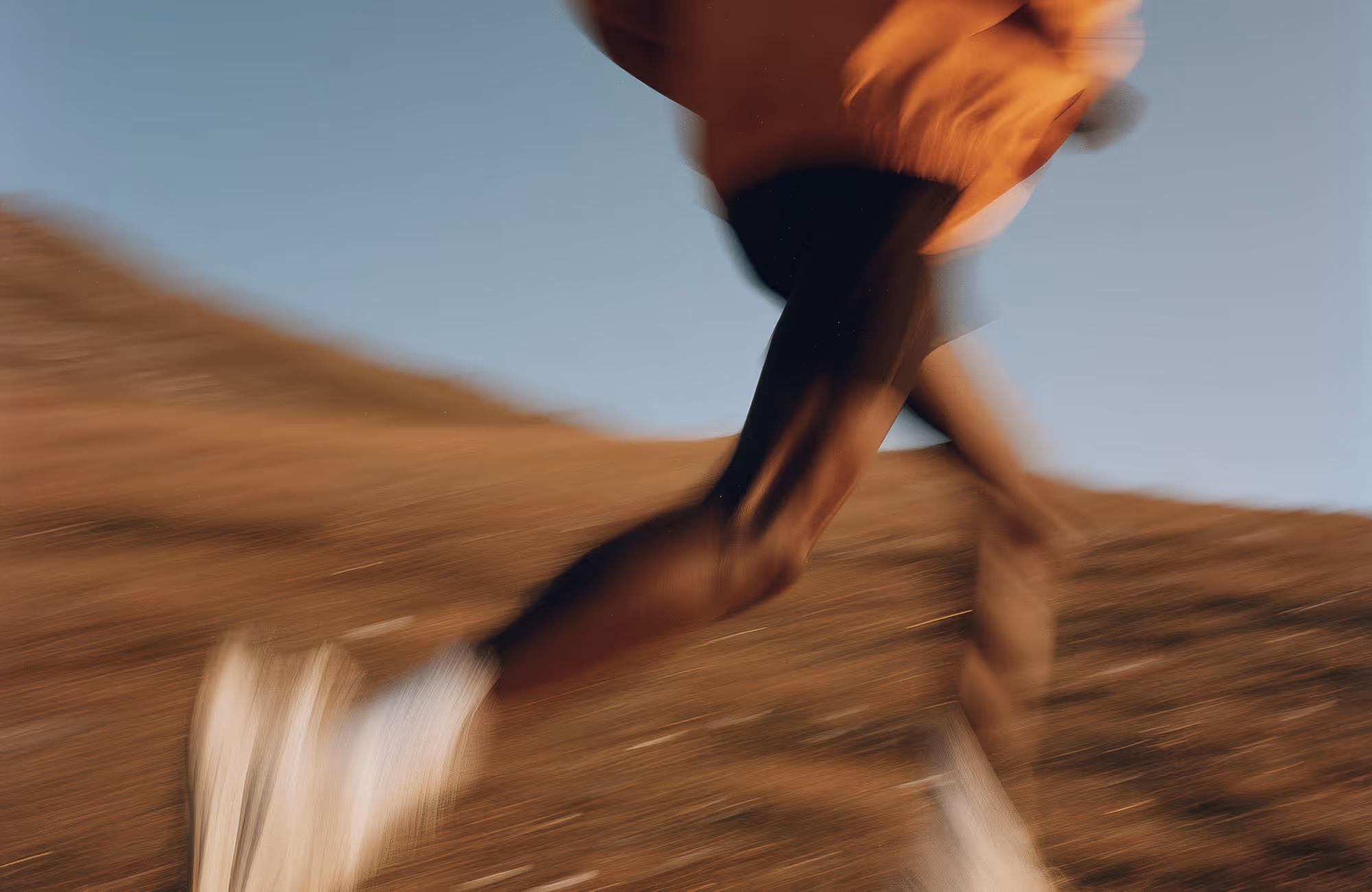 Blurred motion of a runner's muscular legs on a dry hillside trail under a clear blue sky.