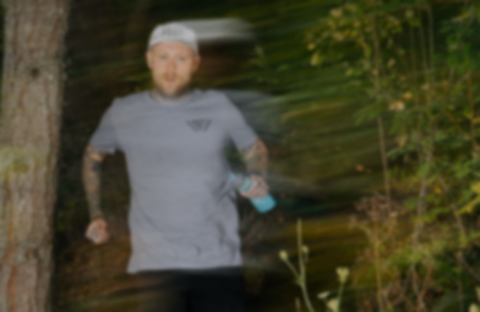 Blurry image of a person in a gray shirt and cap running outdoors near a tree with green foliage in the background.