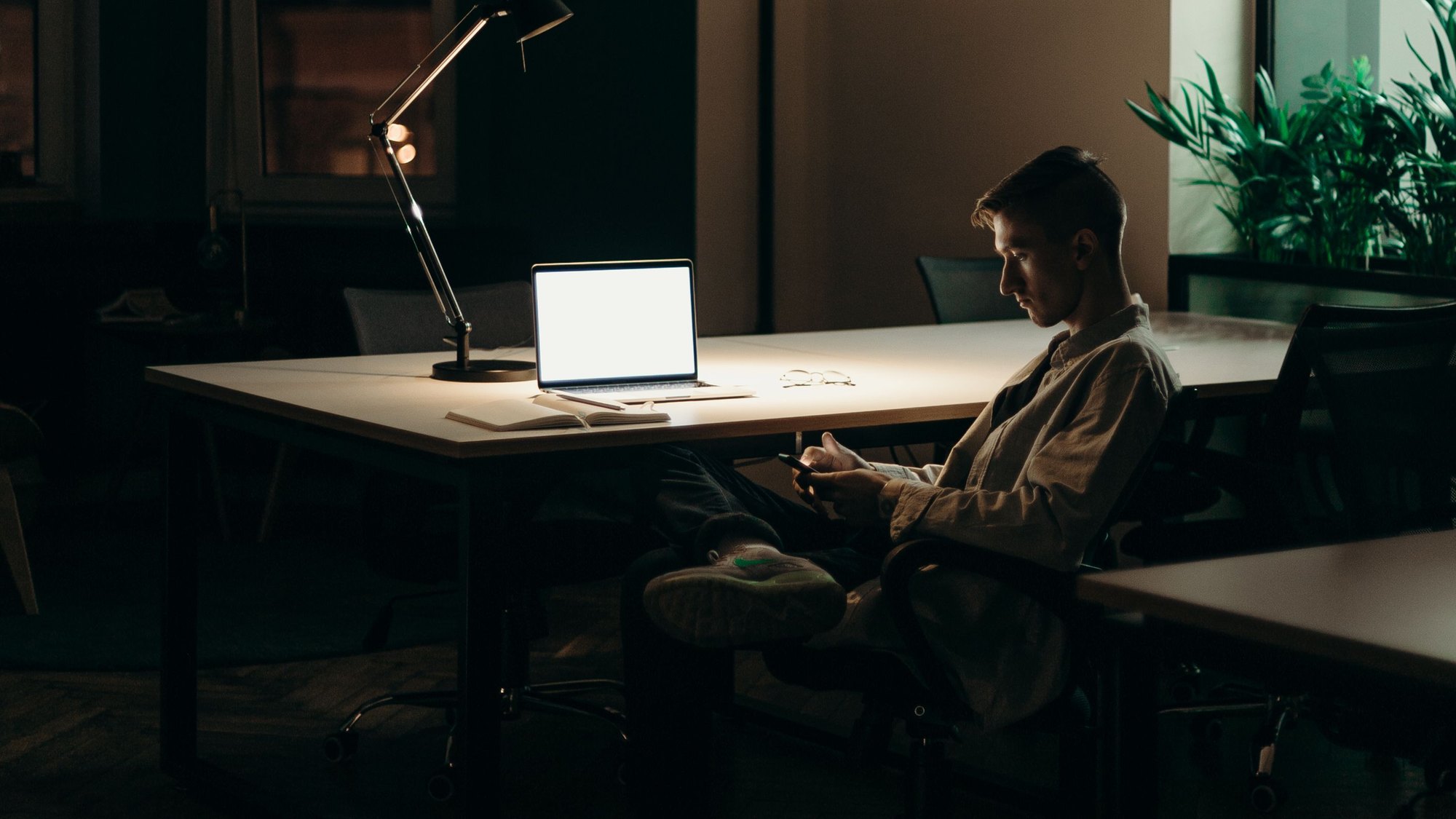 A man alone in a dark office late at night, checking his phone beside a lit desk lamp and laptop.