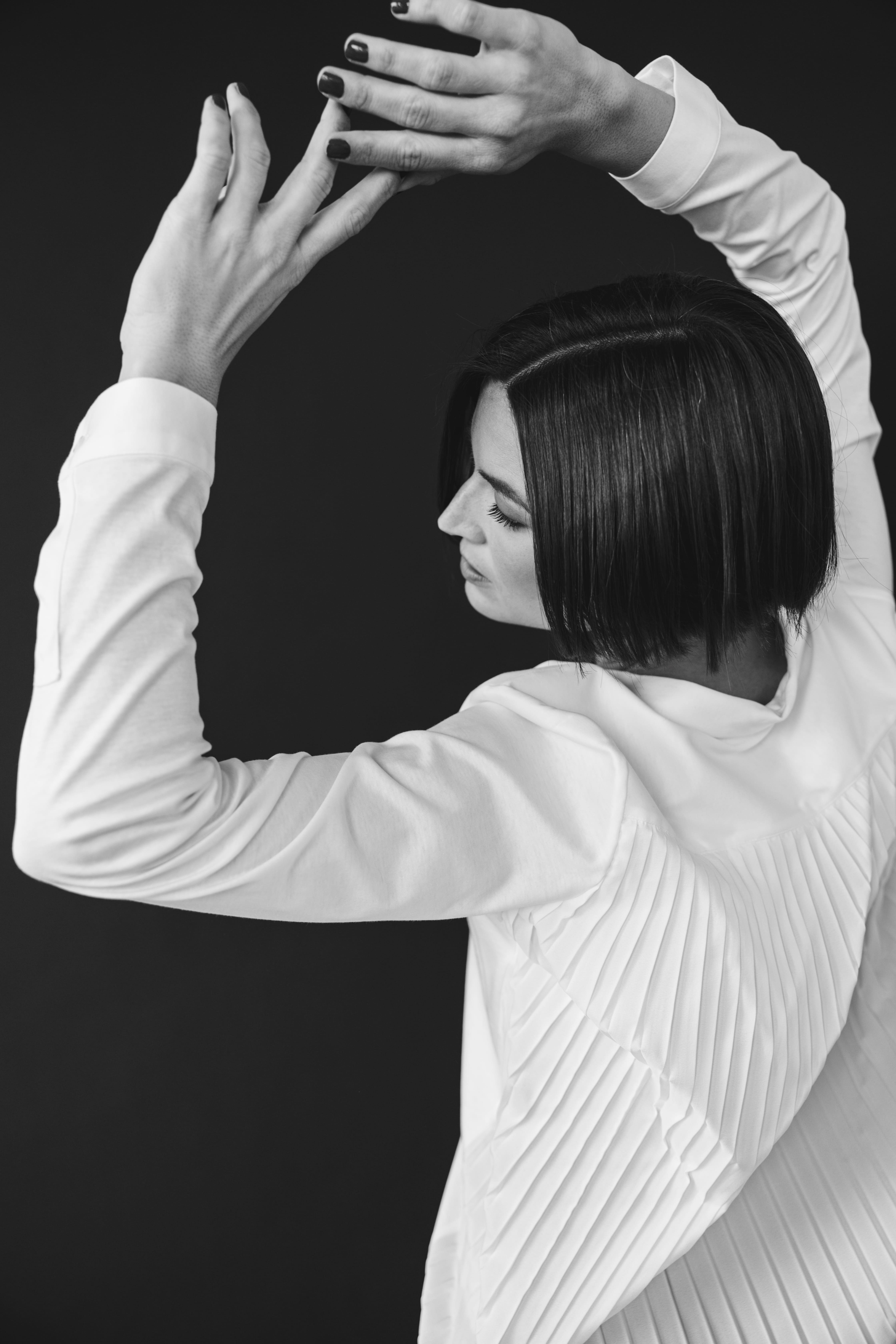 Person with short bob haircut wearing a white pleated-back shirt facing away against a dark background.