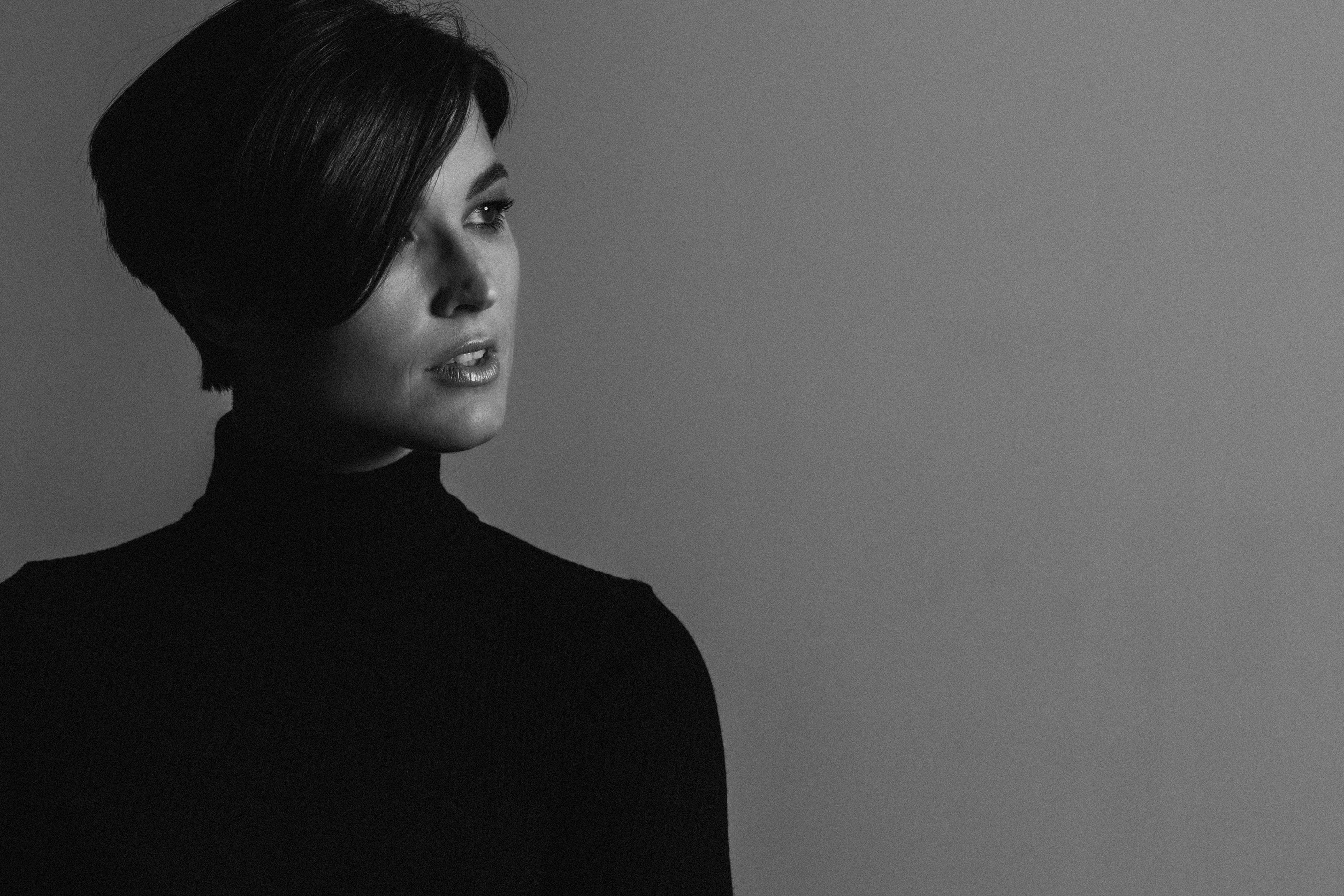 Person with short bob haircut wearing a white pleated-back shirt facing away against a dark background.