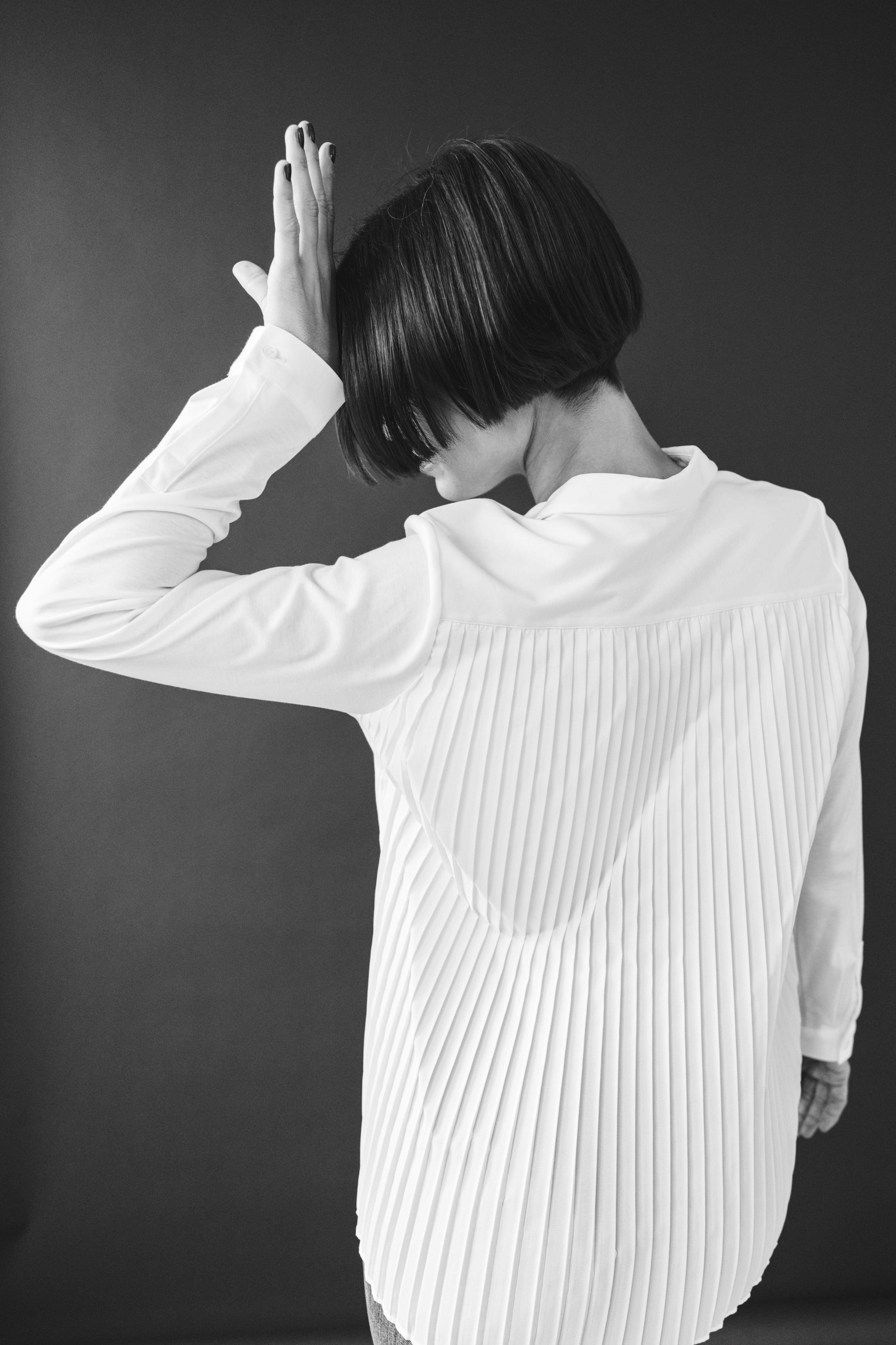 Person with short bob haircut wearing a white pleated-back shirt facing away against a dark background.