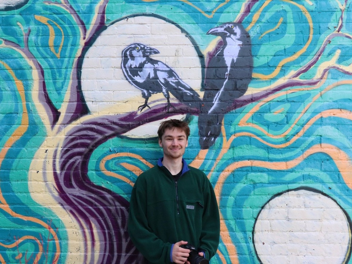 Photo of Owen Smith holding is camera in front of a mural with two crows.