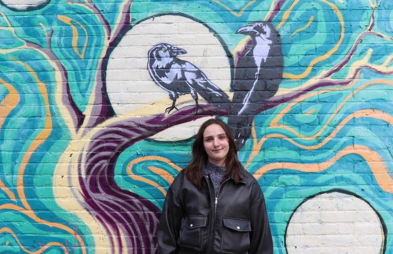Photo of Mackenzie Charbonneau in front of a mural with two crows.
