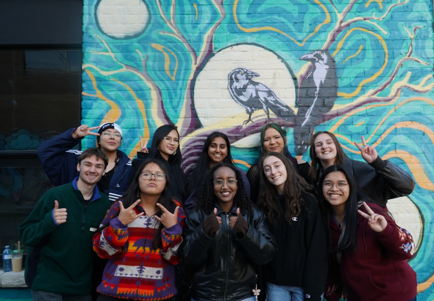 Group photo of Indigenous youth and Carleton students in front of a blue crow mural