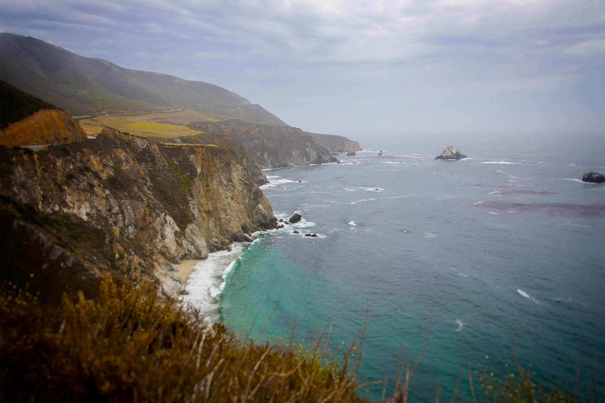 Rocky coastline with cliffs and waves crashing into a small sandy beach beneath a cloudy sky.