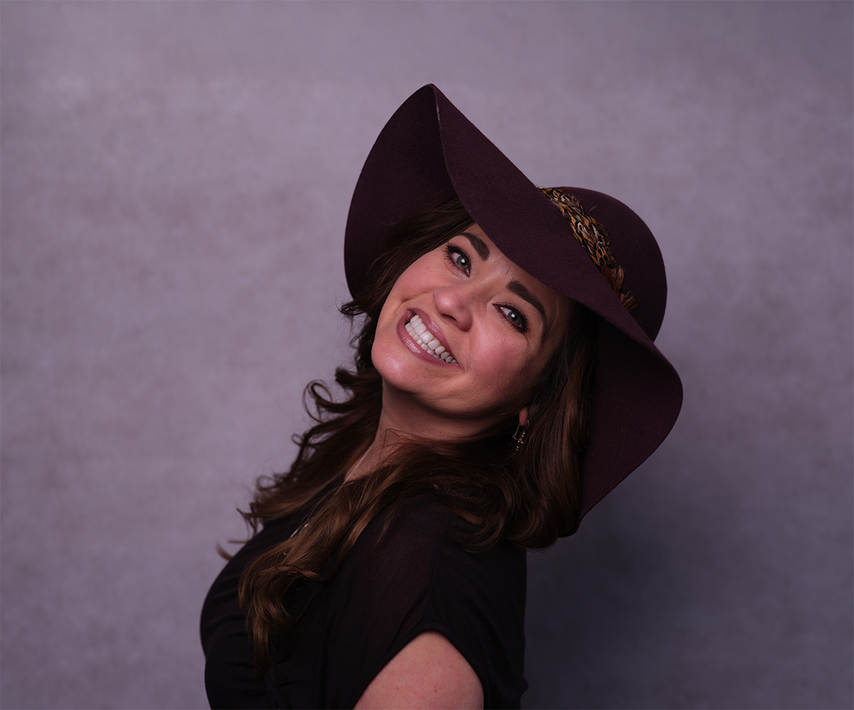 Smiling woman with long brown hair wearing a dark purple wide-brimmed hat against a muted purple background.