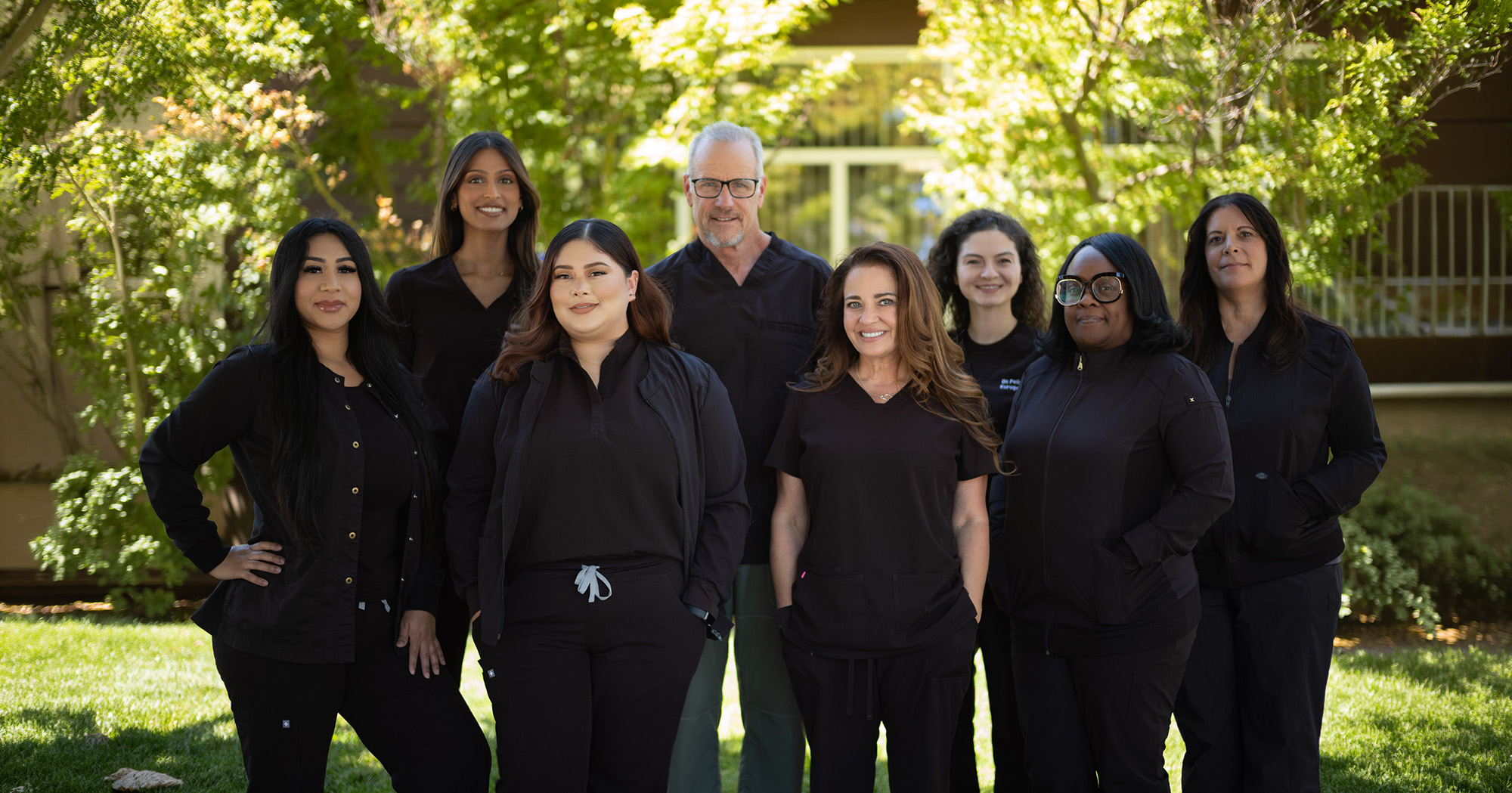 Group portrait of eight healthcare professionals wearing black scrubs standing outside with greenery in the background.