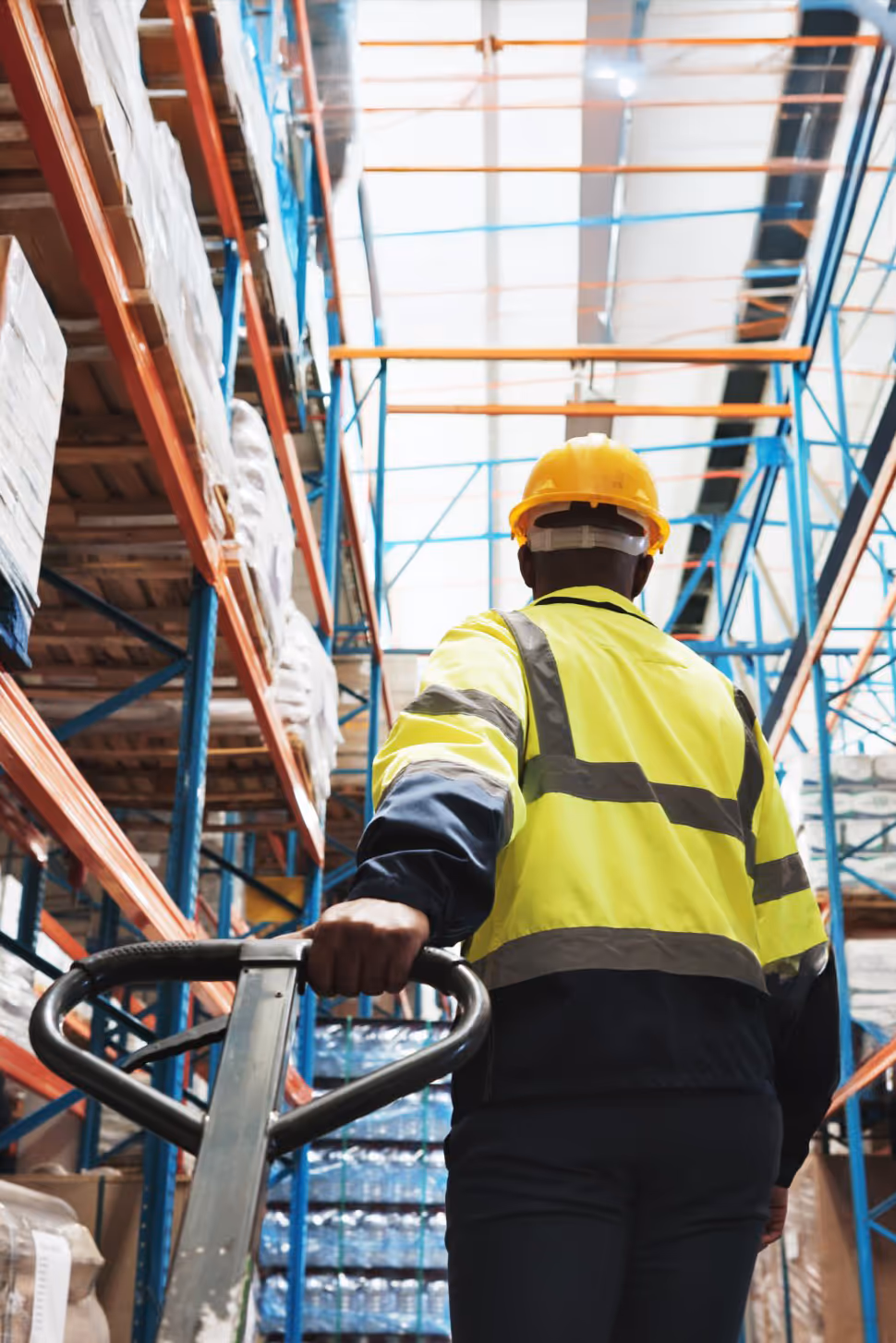 Warehouse worker wearing a yellow hard hat and reflective jacket pushing a pallet jack in an industrial storage area.