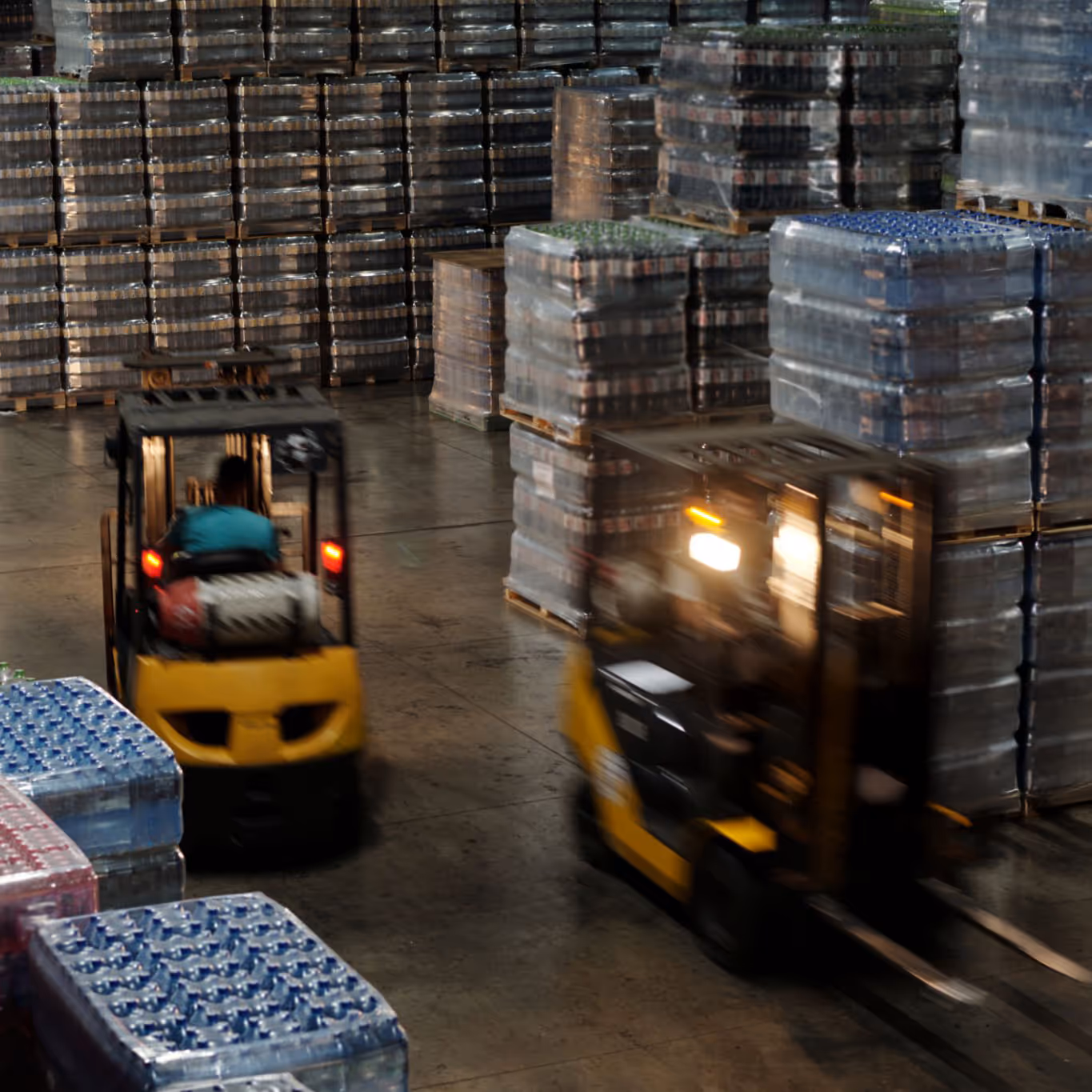 Warehouse interior with two yellow forklifts moving pallets wrapped in plastic.