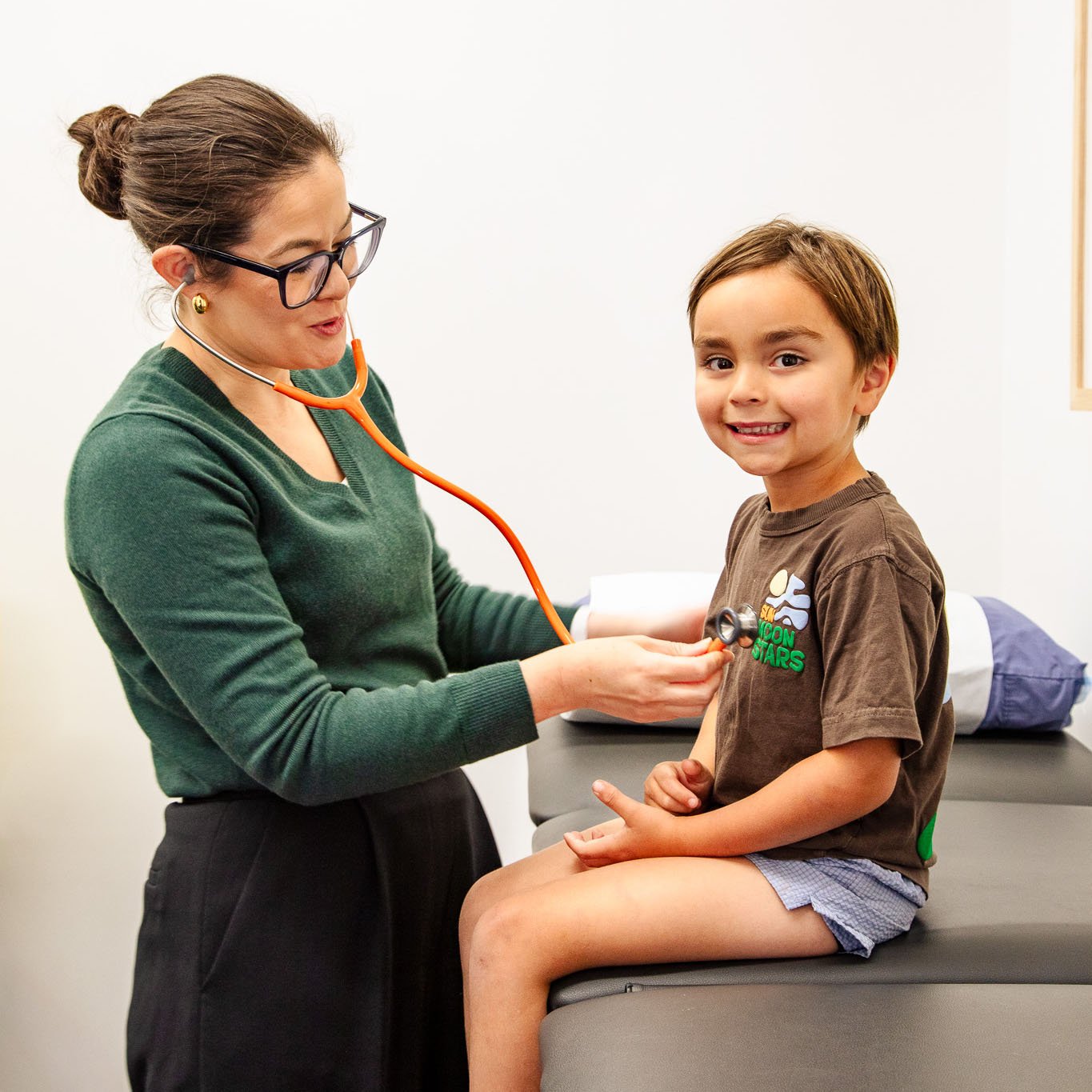 Female doctor using an orange stethoscope to examine a smiling young boy sitting on a medical exam table.