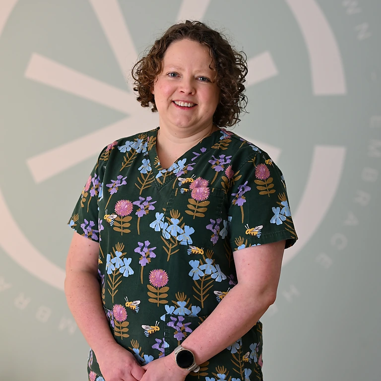 Smiling woman with curly hair wearing a dark green dress and a silver watch, standing against a plain beige background with arms crossed.
