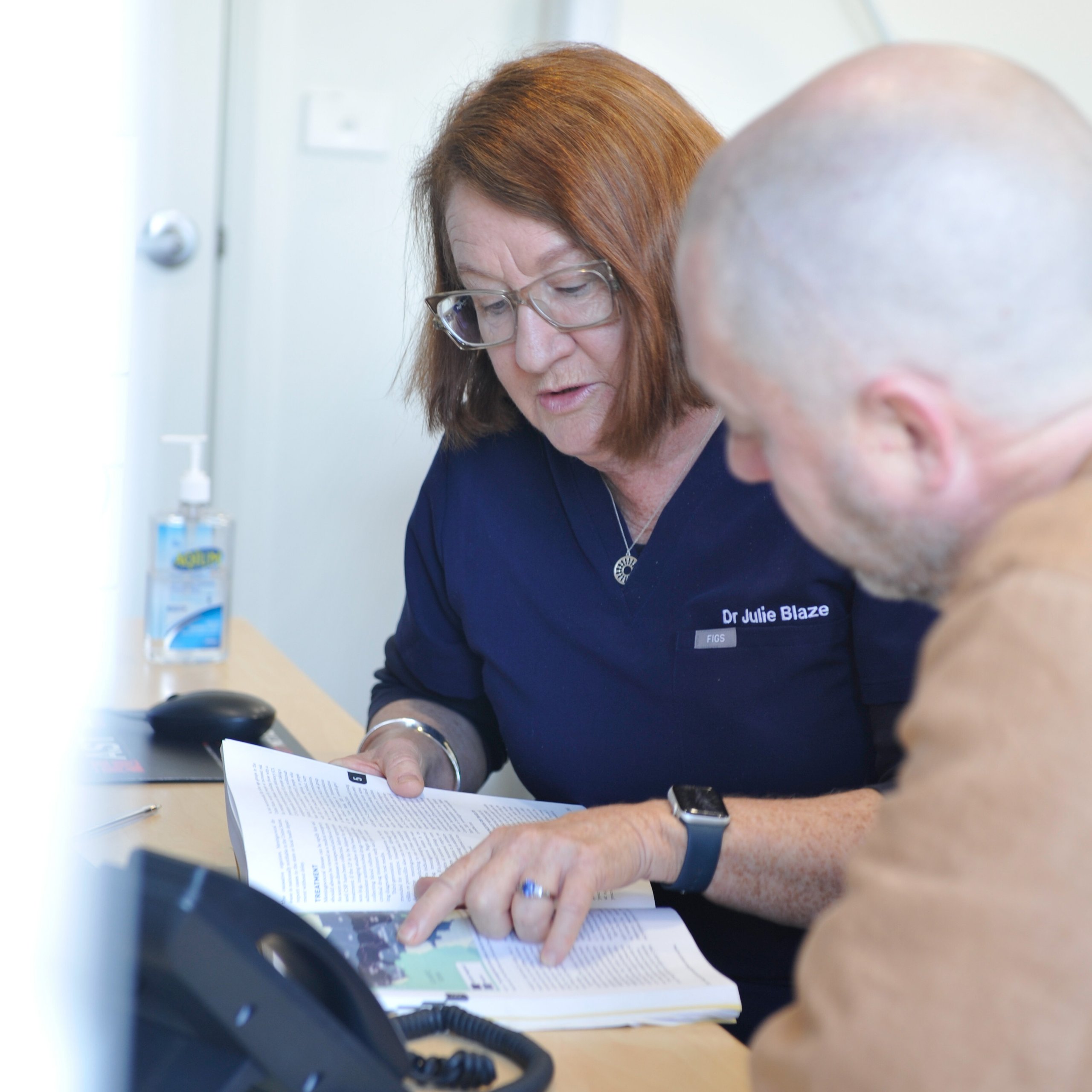 Female doctor with glasses reading and pointing to information in a book while talking to a bald male patient.