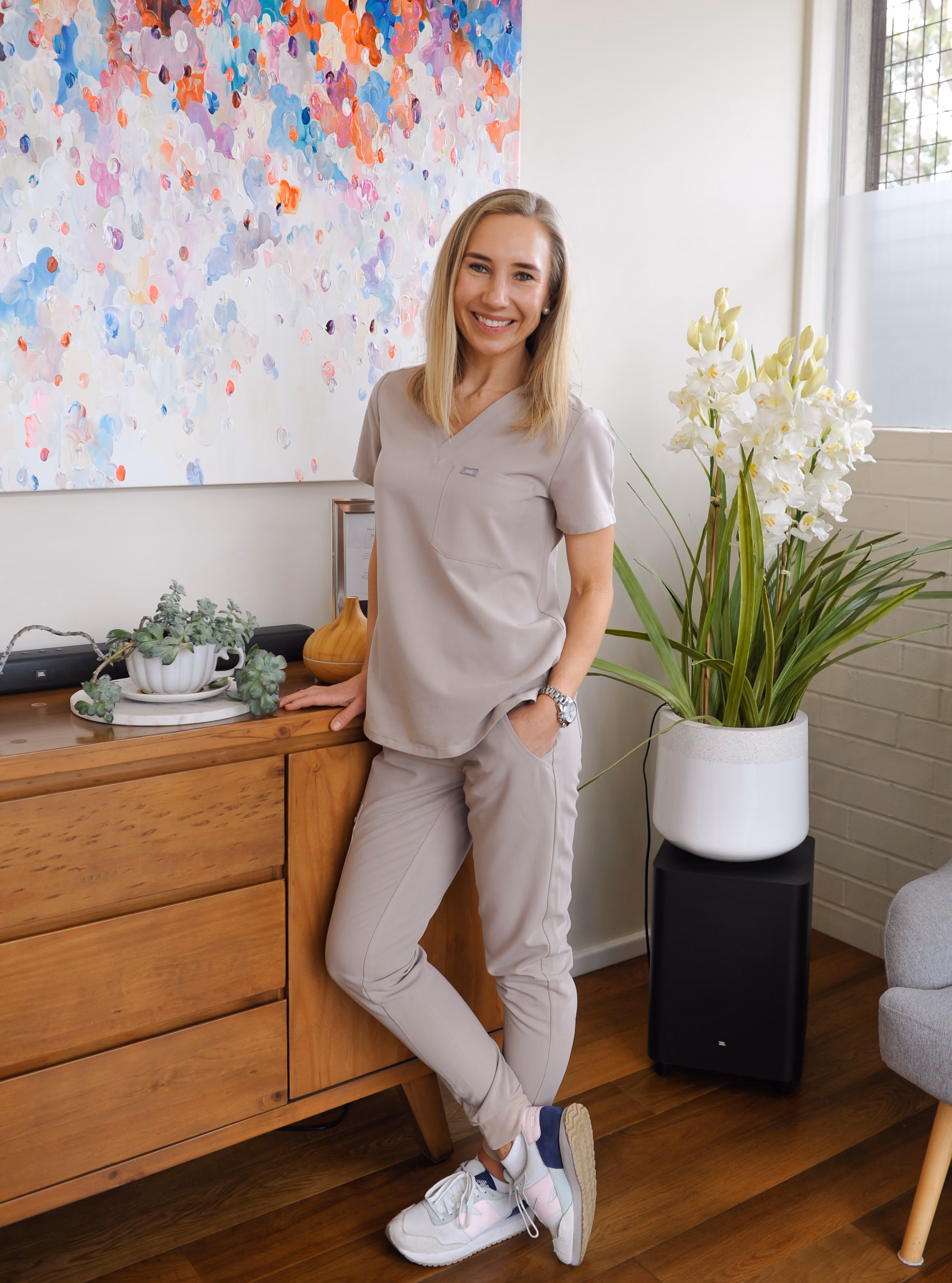 Smiling woman in beige scrubs and sneakers leaning against a wooden cabinet in a bright room with plants and colorful artwork.