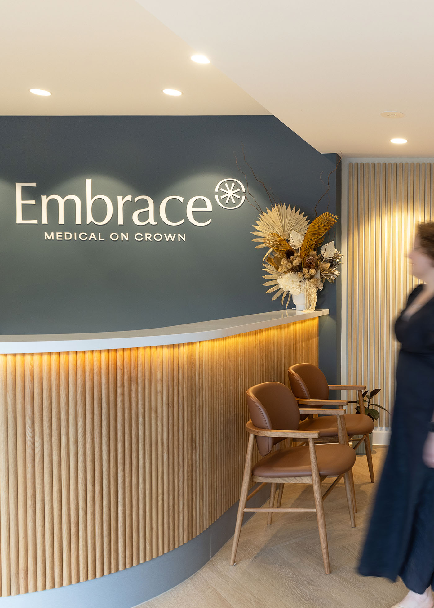 Reception area at Embrace Medical on Crown with wooden slat counter, two brown chairs, and a floral arrangement.