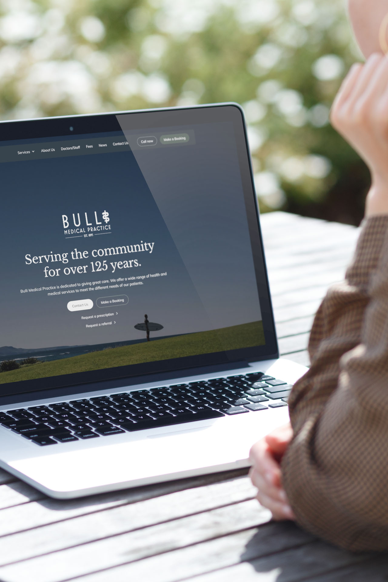 Laptop on a wooden table displaying Bulli Medical Practice homepage with text about serving the community for over 125 years, and a person with folded hands sitting nearby.
