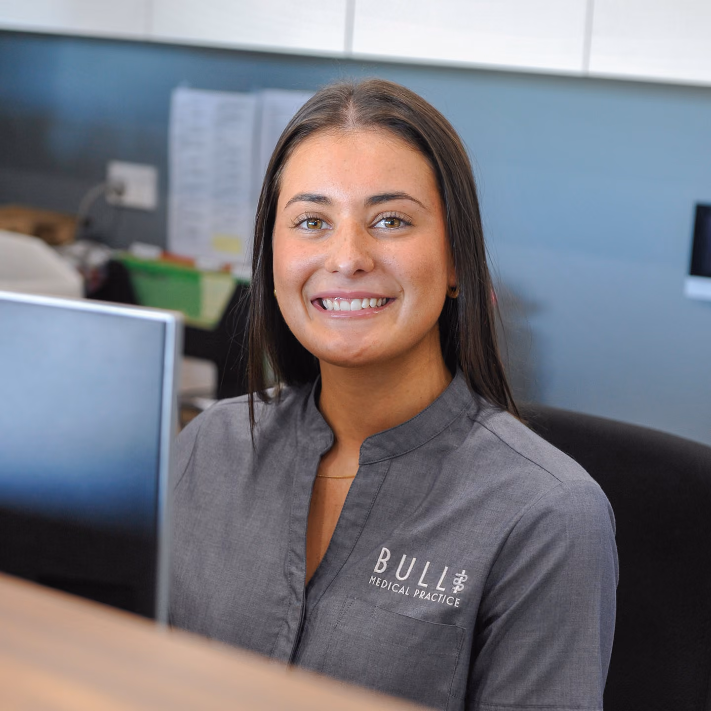 Smiling woman with long brown hair wearing a gray shirt with Bull Medical Practice logo sitting at a desk.