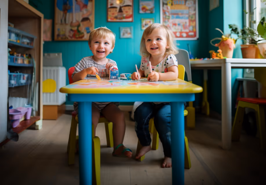 Two young children sitting at a colorful table, smiling and engaged in drawing or coloring activities in a bright room.