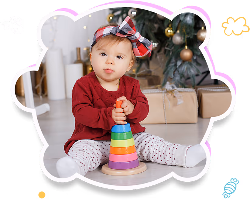 Toddler wearing a red shirt and plaid bow headband sitting on the floor playing with a colorful stacking toy near wrapped Christmas presents.