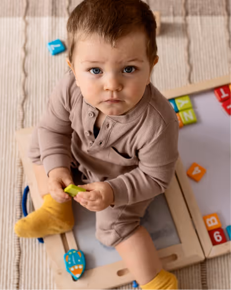 Young child with blue eyes wearing a beige outfit and yellow socks, sitting on a beige rug and playing with colorful letter blocks.