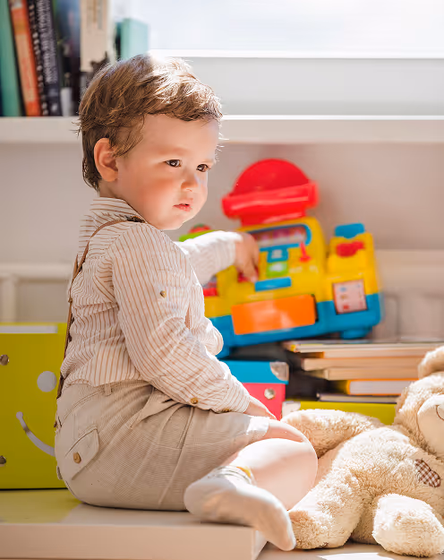 Young child sitting indoors near a large teddy bear and colorful toy, looking thoughtfully to the side.