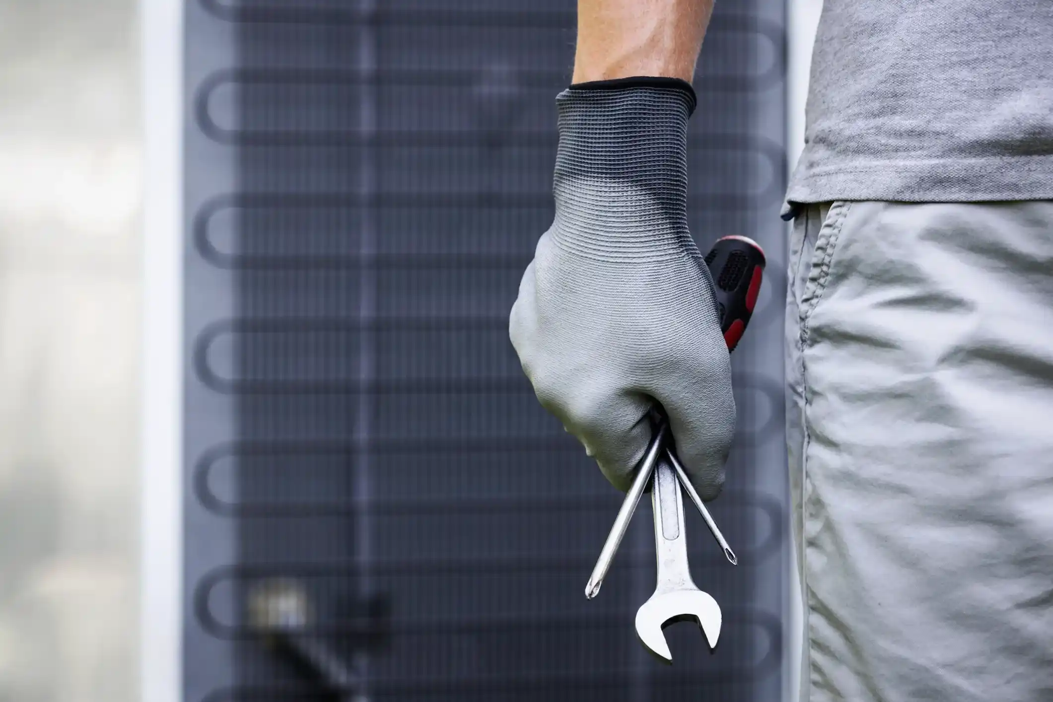Close-up of gloved hand holding wrench, screwdriver, and tool in front of HVAC unit grille, technician wearing gray shirt and shorts.