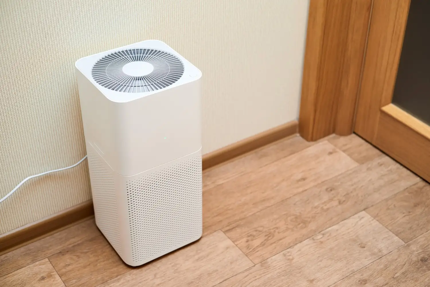 A white, box-shaped air purifier sits on a wooden floor next to a wall and a wooden door frame.