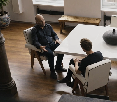 Two men sitting face to face in armchairs having a conversation across a white table in a room with wooden floors.