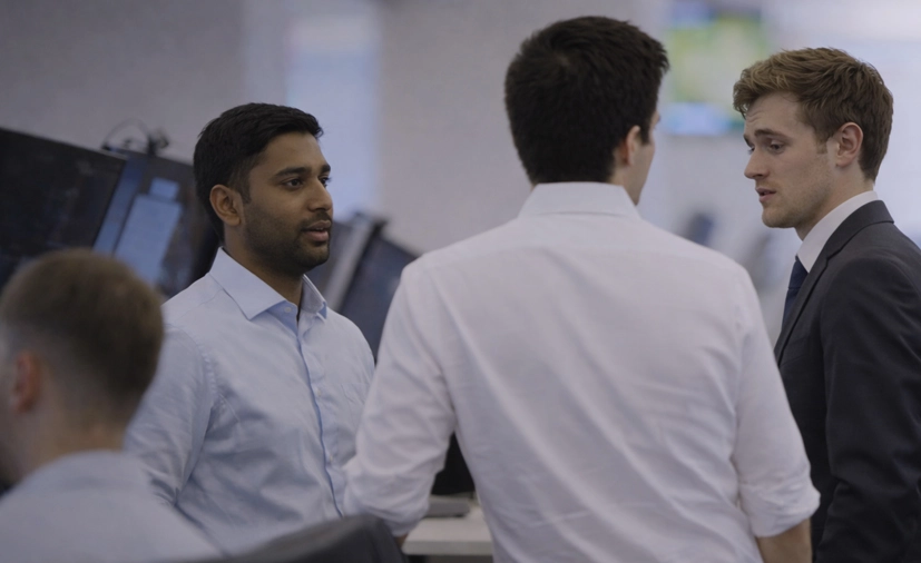 Three men in business attire engaged in a conversation in an office with computer monitors.