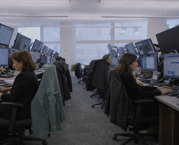 Office workspace with two rows of desks facing each other, multiple monitors on each desk, and several people working on computers.